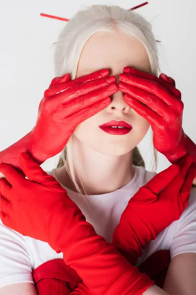 Mãos femininas em tinta vermelha cobrindo os olhos do modelo albino isolado em branco — Fotografia de Stock