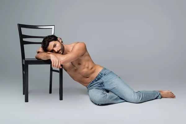 Молодой мужчина без рубашки в джинсах опирается на деревянный стул на сером — стоковое фото