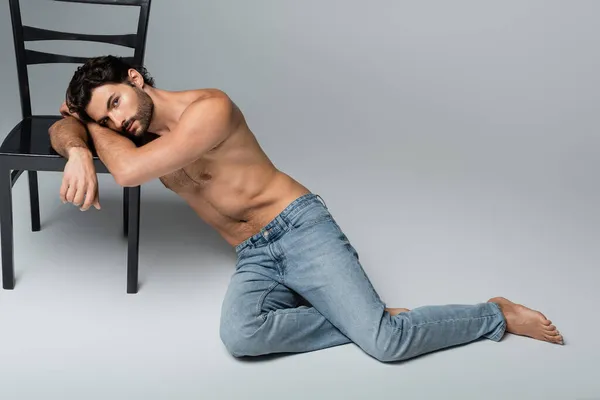 Bearded and shirtless man leaning on wooden chair on grey — Stock Photo