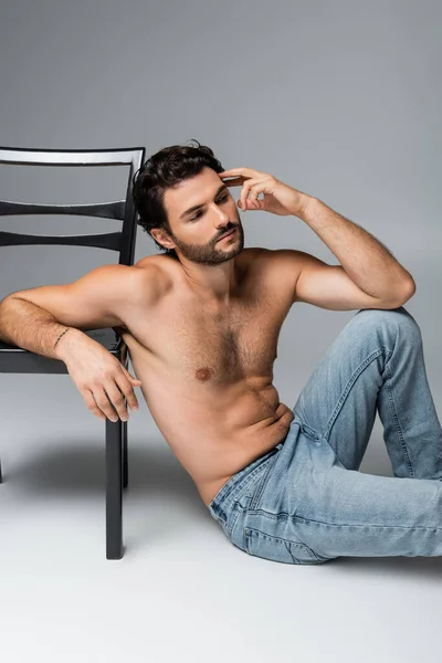 Tattooed and shirtless man in jeans leaning on wooden chair on grey — Stock Photo