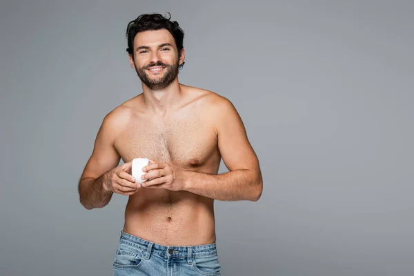 Homem sem camisa e feliz segurando recipiente com creme cosmético isolado em cinza — Fotografia de Stock