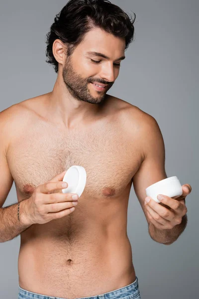 Homem tatuado e sorridente segurando recipiente com creme cosmético isolado em cinza — Fotografia de Stock