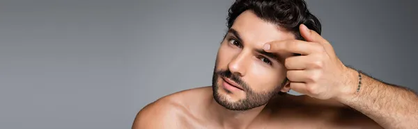 Uomo tatuato e senza maglietta con capelli ondulati applicando crema viso isolata su grigio, banner — Foto stock