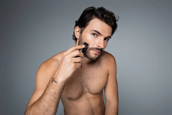 Молодой мужчина без рубашки стрижет бороду изолированный на сером — стоковое фото