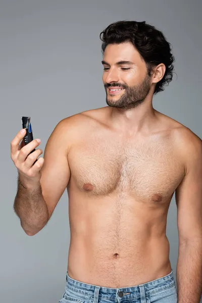 Joven hombre sin camisa sosteniendo maquinilla de afeitar eléctrica y sonriendo aislado en gris - foto de stock