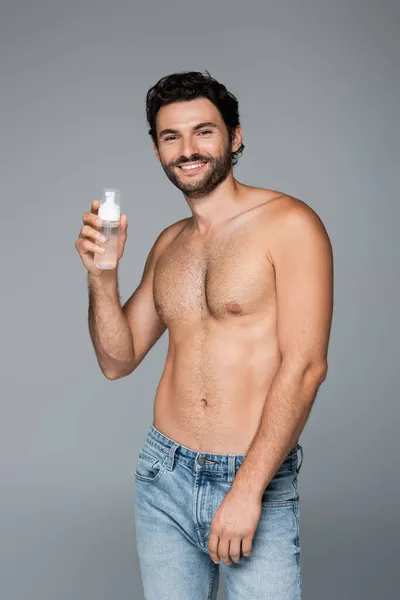 Hombre feliz y sin camisa en jeans con limpiador de espuma aislado en gris - foto de stock