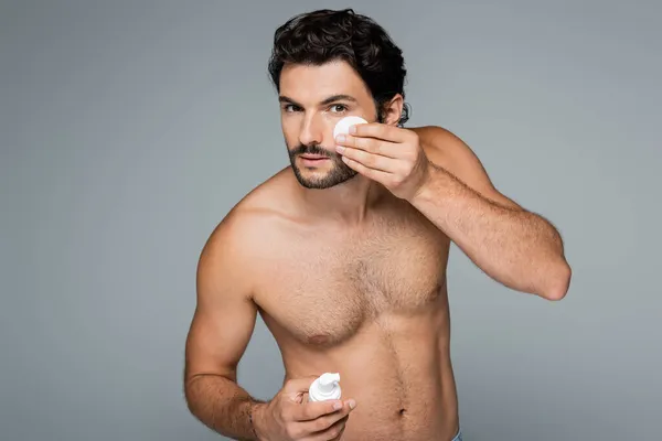 Uomo senza camicia che applica toner sul viso con batuffolo di cotone isolato su grigio — Foto stock