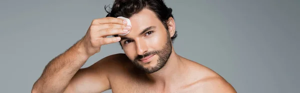 Felice uomo applicando toner sul viso con batuffolo di cotone isolato su grigio, banner — Foto stock