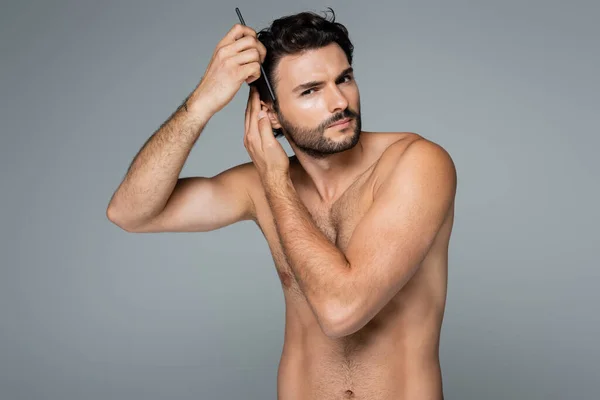 Concentrated shirtless man brushing wavy hair with comb isolated on grey — Stock Photo