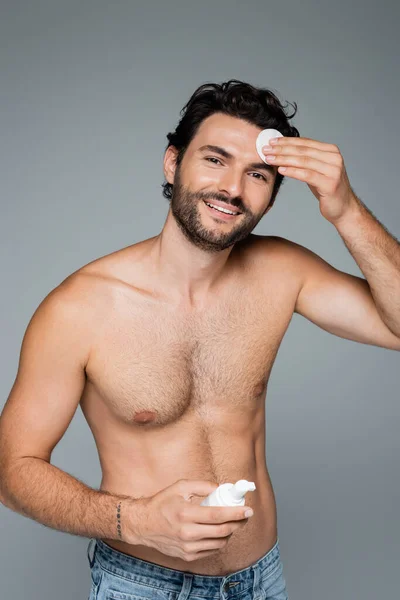 Uomo senza maglietta in jeans sorridente mentre applica toner sul viso con batuffolo di cotone isolato su grigio — Foto stock