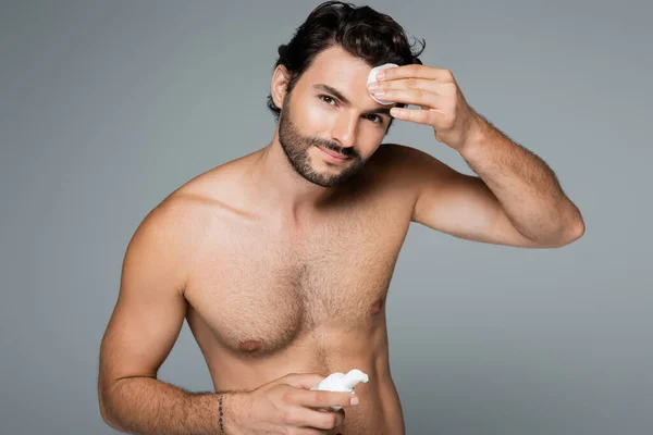 Pleased shirtless man applying toner on face with cotton pad isolated on grey — Stock Photo