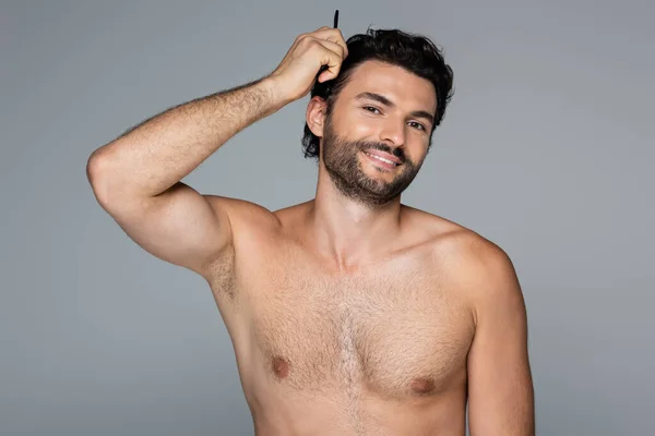 Hombre feliz sin camisa cepillado pelo ondulado con peine aislado en gris - foto de stock