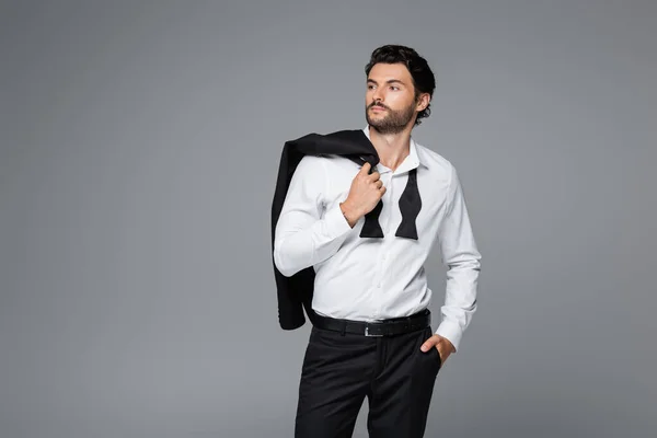 Homme barbu en chemise blanche tenant blazer et debout avec la main dans la poche isolé sur gris — Photo de stock