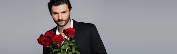 Homme barbu en costume tenant bouquet de roses rouges tout en regardant la caméra isolée sur gris, bannière — Photo de stock