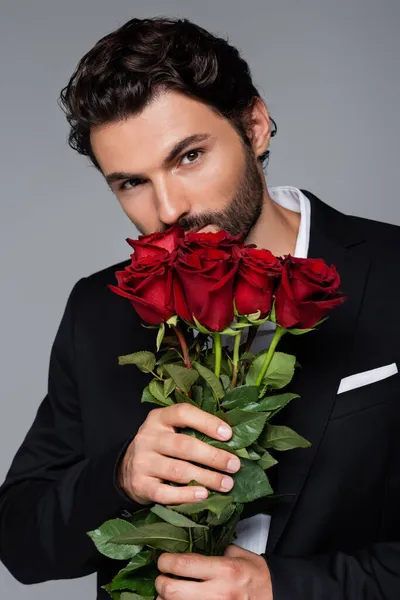 Hombre barbudo en desgaste formal oliendo rosas rojas mientras mira la cámara aislada en gris - foto de stock