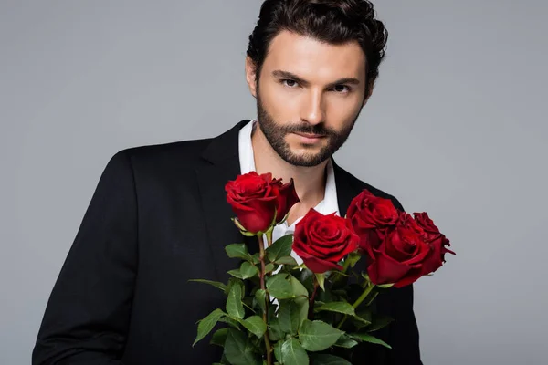 Bearded young man in suit holding bouquet of red roses while looking at camera isolated on grey — Stock Photo