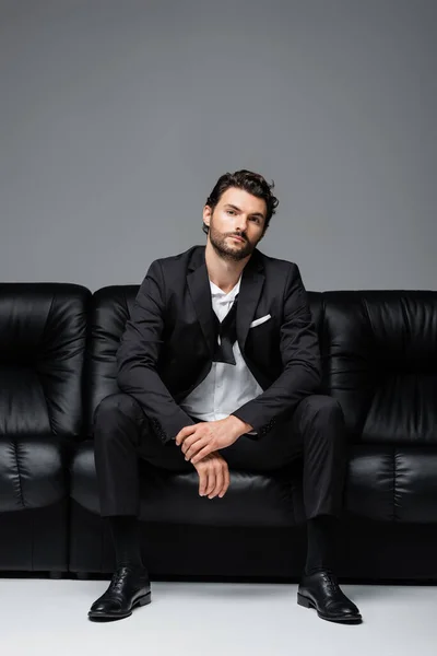Bearded man in suit sitting on black leather sofa on grey — Stock Photo