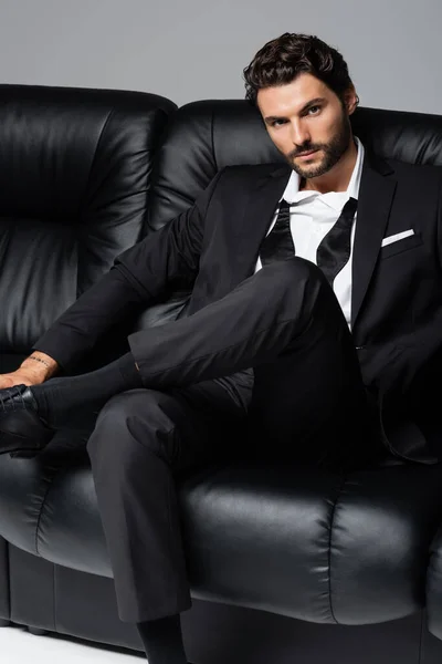 Татуированный и бородатый мужчина в формальной одежде сидит на черном диване, изолированном на сером — стоковое фото