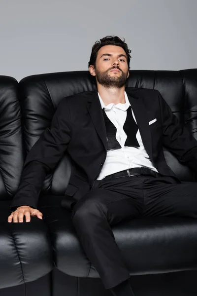 Joven barbudo en ropa formal sentado en sofá negro aislado en gris - foto de stock