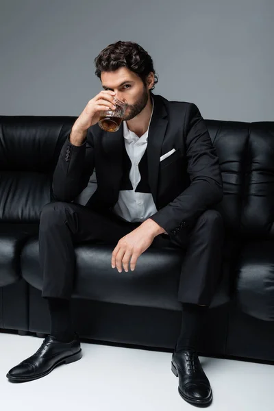 Young bearded man in formal wear sitting on black sofa and drinking whiskey on grey — Stock Photo