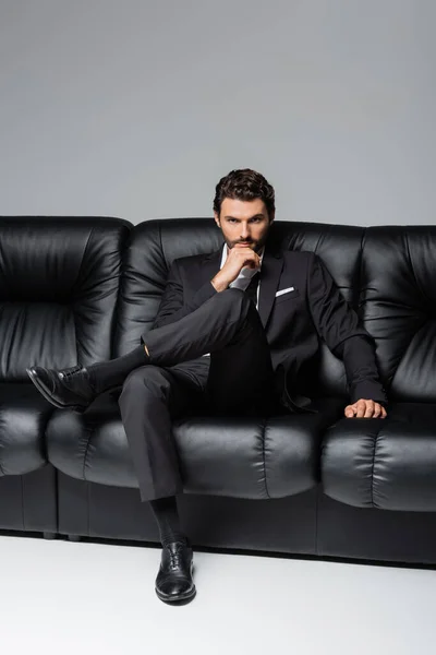 Uomo sicuro e barbuto in abito formale seduto sul divano nero sul grigio — Foto stock