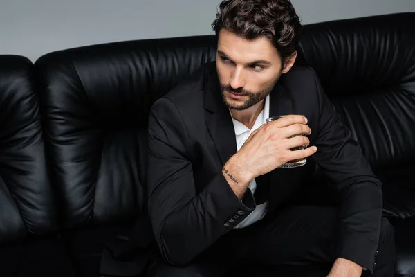 Homme tatoué en tenue de cérémonie assis sur un canapé noir et tenant un verre de whisky isolé sur gris — Photo de stock