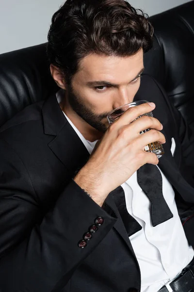 Man in formal wear sitting on black sofa and drinking whiskey isolated on grey — Stock Photo