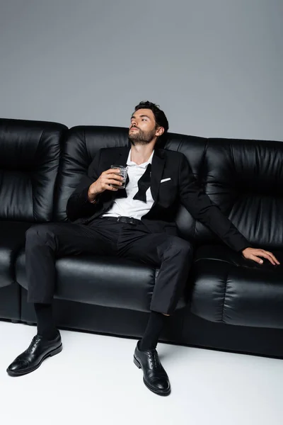 Homem relaxado no desgaste formal sentado no sofá preto e segurando um copo de uísque no cinza — Fotografia de Stock