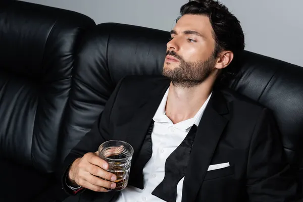Entspannter Mann im Anzug sitzt auf schwarzem Sofa und hält Glas Whisky isoliert auf grau — Stockfoto
