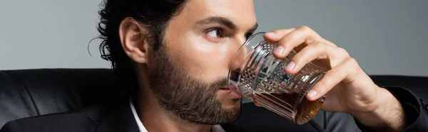 Bärtiger Mann mit welligem Haar trinkt Whisky isoliert auf grau, Banner — Stockfoto