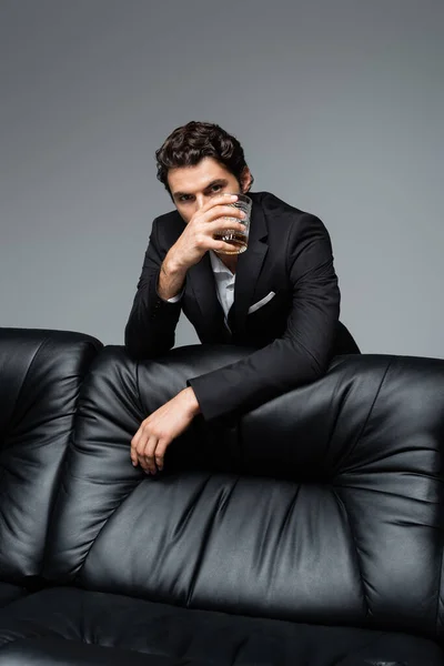 Man in suit standing near black sofa and holding glass of whiskey isolated on grey — Stock Photo