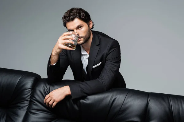 Hombre barbudo en traje de pie cerca de sofá negro y sosteniendo vaso de whisky aislado en gris - foto de stock