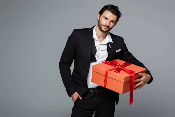 Hombre barbudo de traje posando con caja de regalo roja y de pie con la mano en el bolsillo aislado en gris - foto de stock