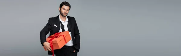 Hombre barbudo en traje posando con caja de regalo roja y de pie con la mano en el bolsillo aislado en gris, bandera - foto de stock