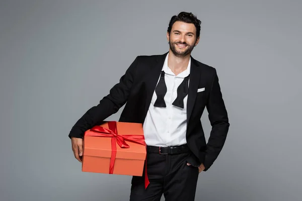 Joyful man in suit holding red gift box and standing with hand in pocket isolated on grey — Stock Photo