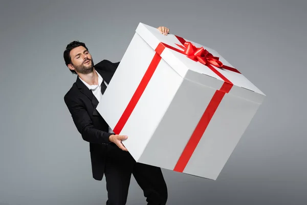 Бородатий чоловік у костюмі тримає величезну подарункову коробку ізольовано на сірому — стокове фото