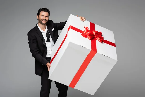 Веселий чоловік у костюмі тримає величезну подарункову коробку ізольовано на сірому — стокове фото