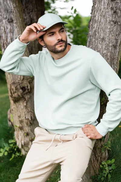 Bearded man adjusting cap and standing near tree trunks in park — Stock Photo