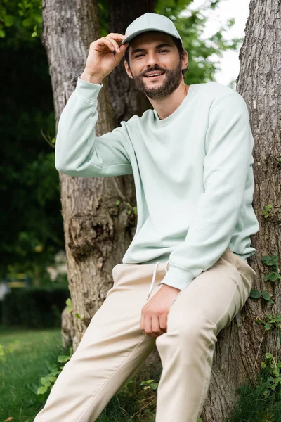 Cheerful man adjusting cap and leaning on tree trunks in park — Stock Photo