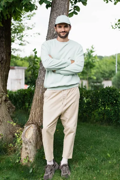 Vollbärtiger Mann mit Mütze steht mit verschränkten Armen im Park — Stockfoto