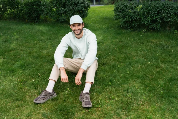 Comprimento total do homem barbudo na tampa sorrindo enquanto sentado no gramado verde — Fotografia de Stock