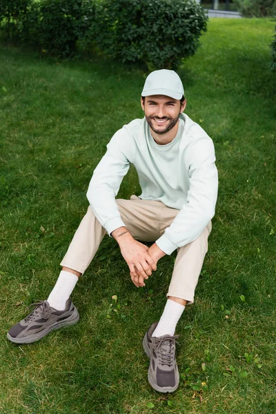 Piena lunghezza di uomo soddisfatto in berretto sorridente mentre seduto sul prato verde — Foto stock