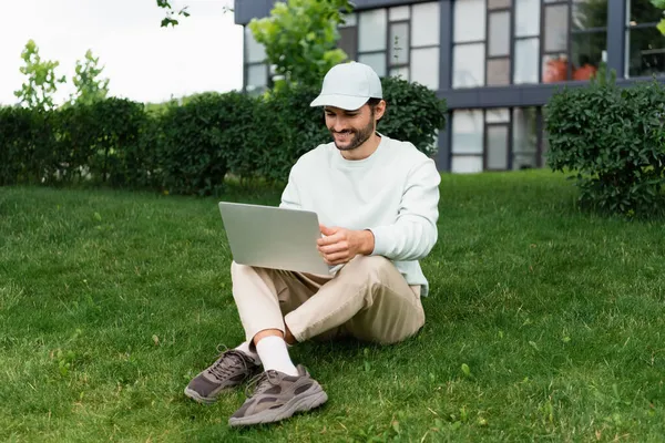 Longitud completa de freelancer feliz usando el ordenador portátil mientras está sentado con las piernas cruzadas en el césped - foto de stock