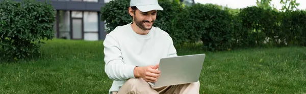 Full length of happy freelancer using laptop while sitting on lawn, banner — Stock Photo