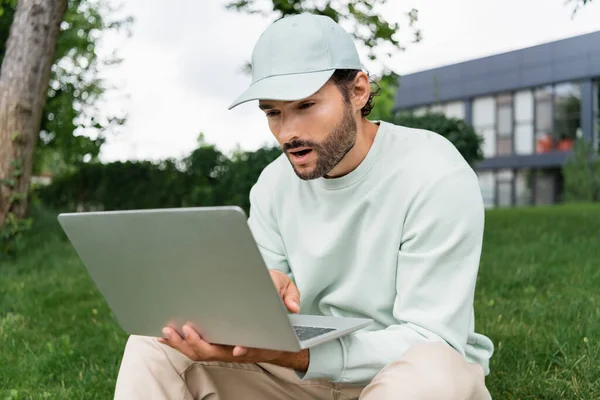 Freelancer chocado em cap olhando para laptop no parque — Fotografia de Stock