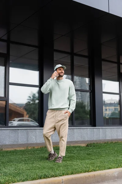 Pleine longueur de l'homme heureux debout avec la main dans la poche et parler sur le téléphone portable près du bâtiment moderne — Photo de stock