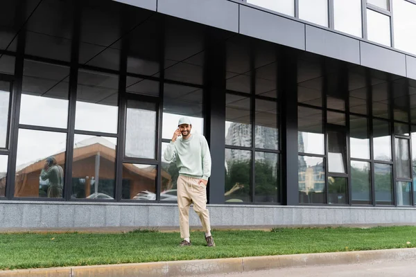 Full length of cheerful man standing with hand in pocket and talking on cellphone near modern building — Stock Photo