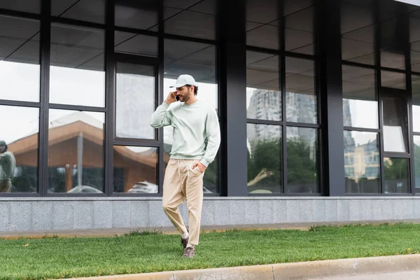 Junger Mann mit Schirmmütze steht mit der Hand in der Tasche und telefoniert in der Nähe moderner Gebäude — Stockfoto