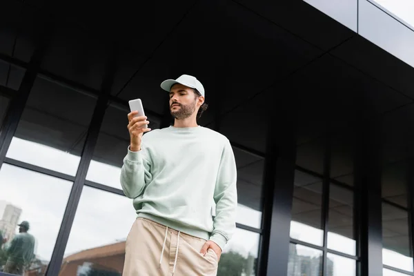 Tiefansicht eines jungen Mannes mit Mütze, der mit der Hand in der Tasche in der Nähe moderner Gebäude steht und Smartphone benutzt — Stockfoto