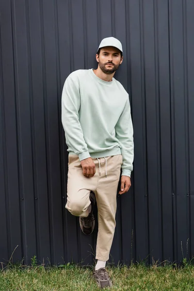 Full length of bearded man in baseball cap posing near metallic fence — Stock Photo